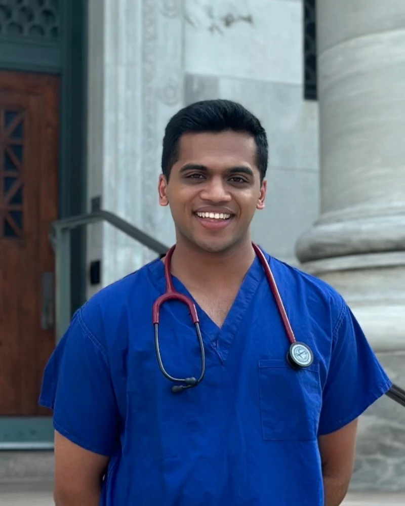 Neil Sardesai smiling in blue scrubs with a stethoscope, standing on the steps of Gordon Hall